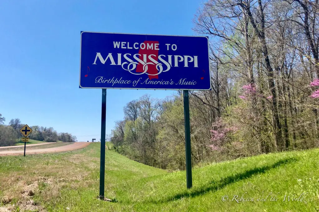 "Welcome to Mississippi" sign by the roadside, proclaiming Mississippi as the Birthplace of America's Music, with a clear blue sky and lush greenery in the background. The Visit Mississippi welcome sign that you'll see when you enter the state.