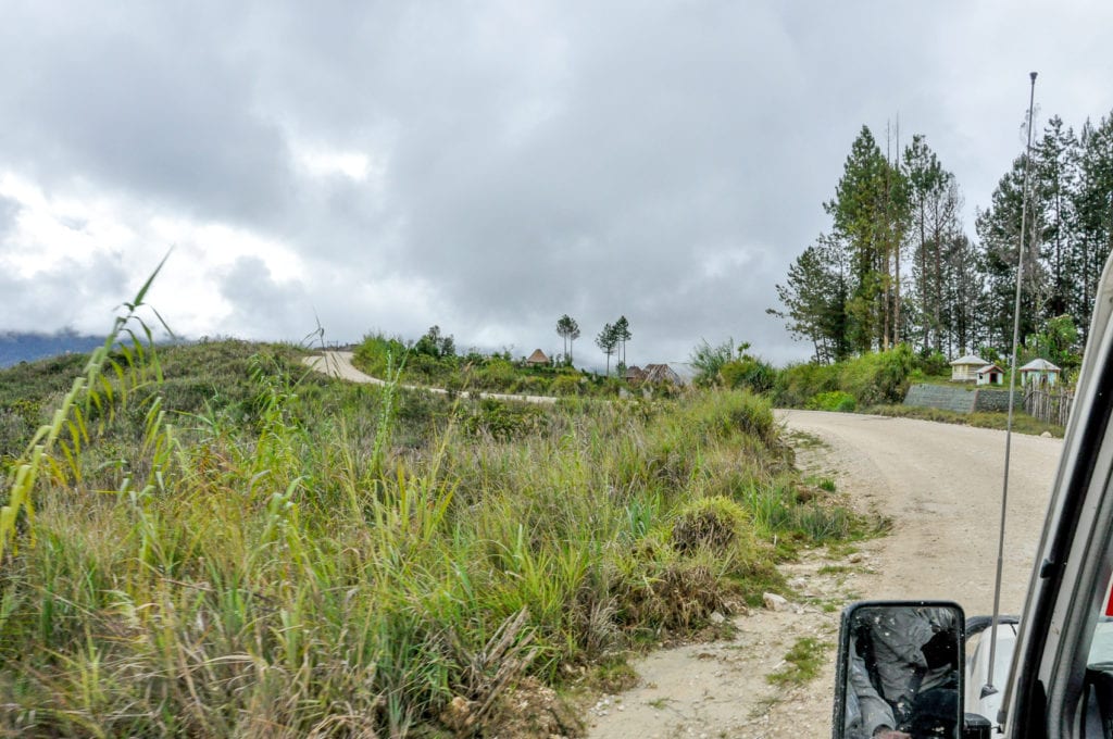 Rural road through a grassy landscape with scattered trees and houses, illustrating a typical drive in the highlands of Papua New Guinea. A portion of the Highlands Highway, one of the few major roads in PNG.