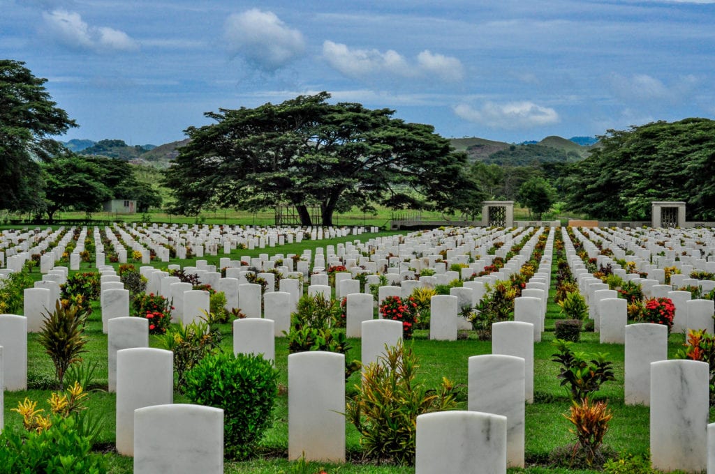 Well-maintained war cemetery with rows of white grave markers, lush green grass and flowering plants under a cloudy sky. Bomana Cemetery, near Port Moresby, is the resting place of almost 4,000 soldiers.