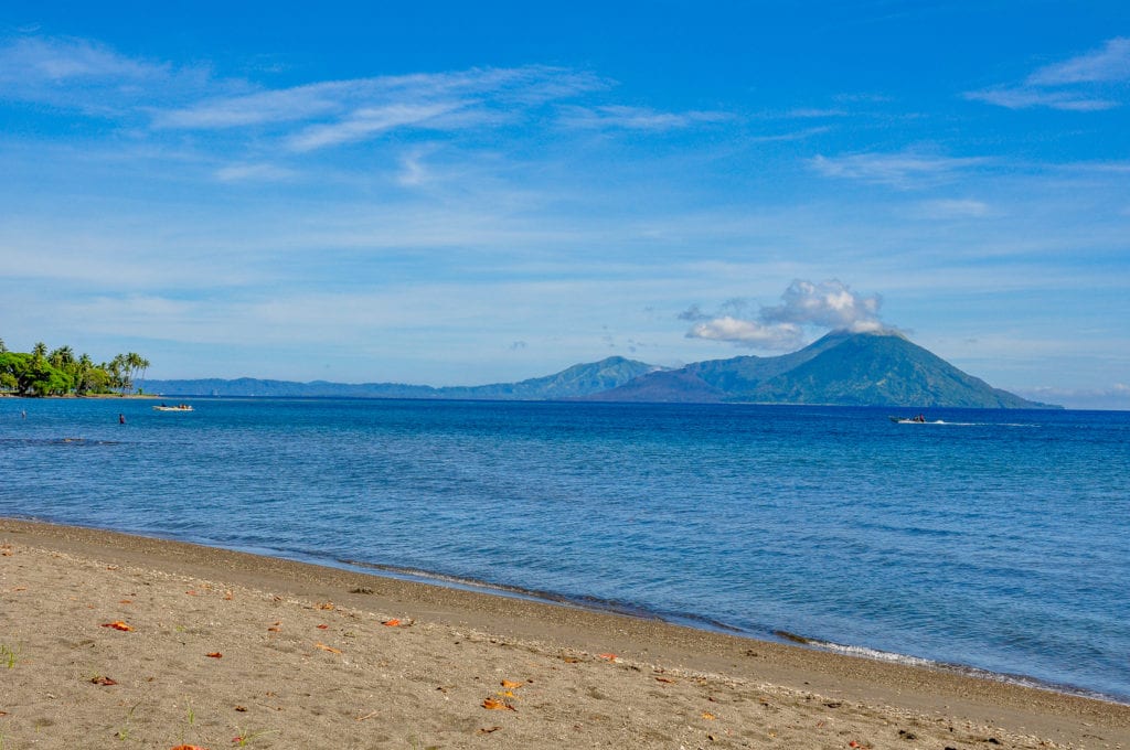 A serene seaside view in Papua New Guinea, showcasing a calm beach with a distant volcano and clear blue skies. There are almost 100 volcanoes in PNG.