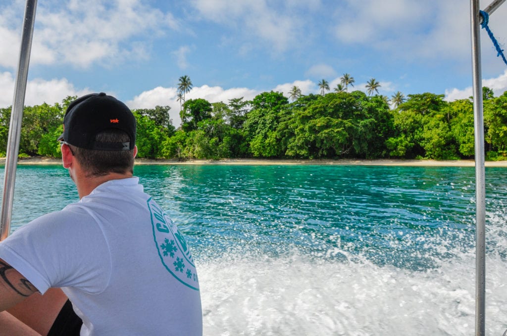 A man - the author's husband - sitting on the edge of a boat looking at a tropical island. There are so many stunning beaches and coastal areas to explore in PNG.
