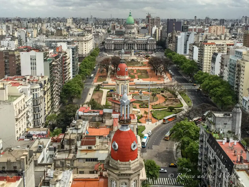 An aerial view of Buenos Aires with dense, varied architecture, a large green park in the center, and a striking red-domed building in the foreground. The background shows a cloudy sky over the urban expanse. There are great views of Buenos Aires from the top of Palacio Barolo.