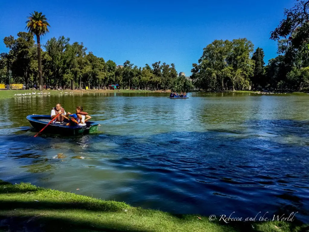 Two people rowing a boat on a calm lake in a park, with other boaters in the background and lush greenery surrounding the water. Palermo in Buenos Aires is filled with lush green parks and lakes that are fun to visit on the weekend - it's a great Buenos Aires neighbourhood to stay in.
