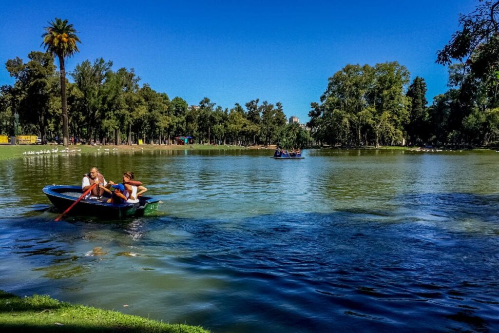 Two people rowing a boat on a calm lake in a park, with other boaters in the background and lush greenery surrounding the water. Palermo in Buenos Aires is filled with lush green parks and lakes that are fun to visit on the weekend - it's a great Buenos Aires neighbourhood to stay in.