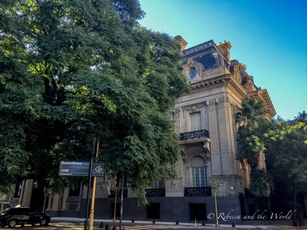 A historic building with elaborate stone facade and balconies, shaded by tall green trees, on a quiet Buenos Aires street corner. This is in the Recoleta neighbourhood.