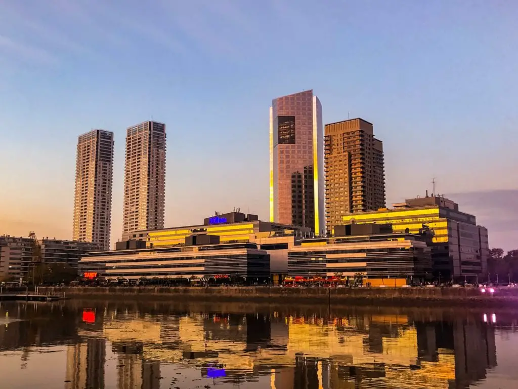 The Buenos Aires skyline during sunset reflecting on the water, with tall buildings illuminated by the warm hues of the setting sun. Puerto Madero is one of Buenos Aires neighbourhoods.
