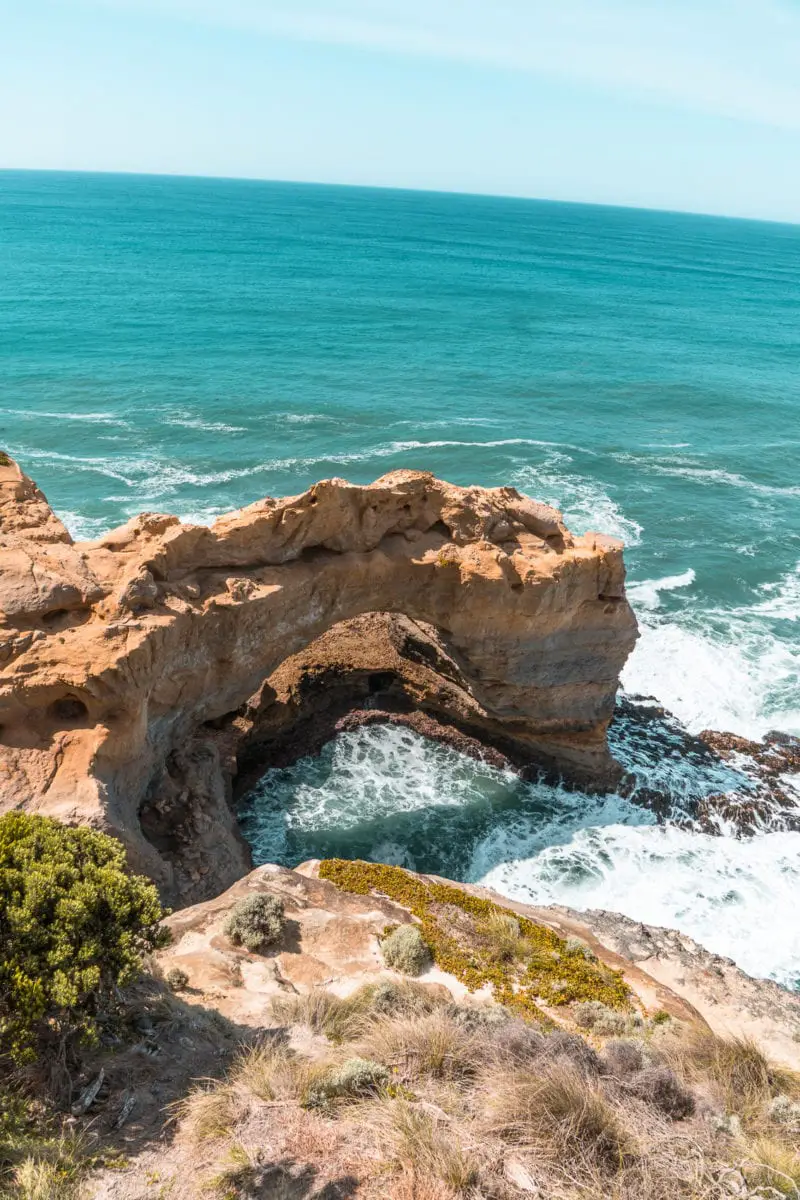 Rock archway called The Arch, with ocean water flowing through it, set against a backdrop of eroded cliffs and blue sea. The Arch is located along the Great Ocean Road.