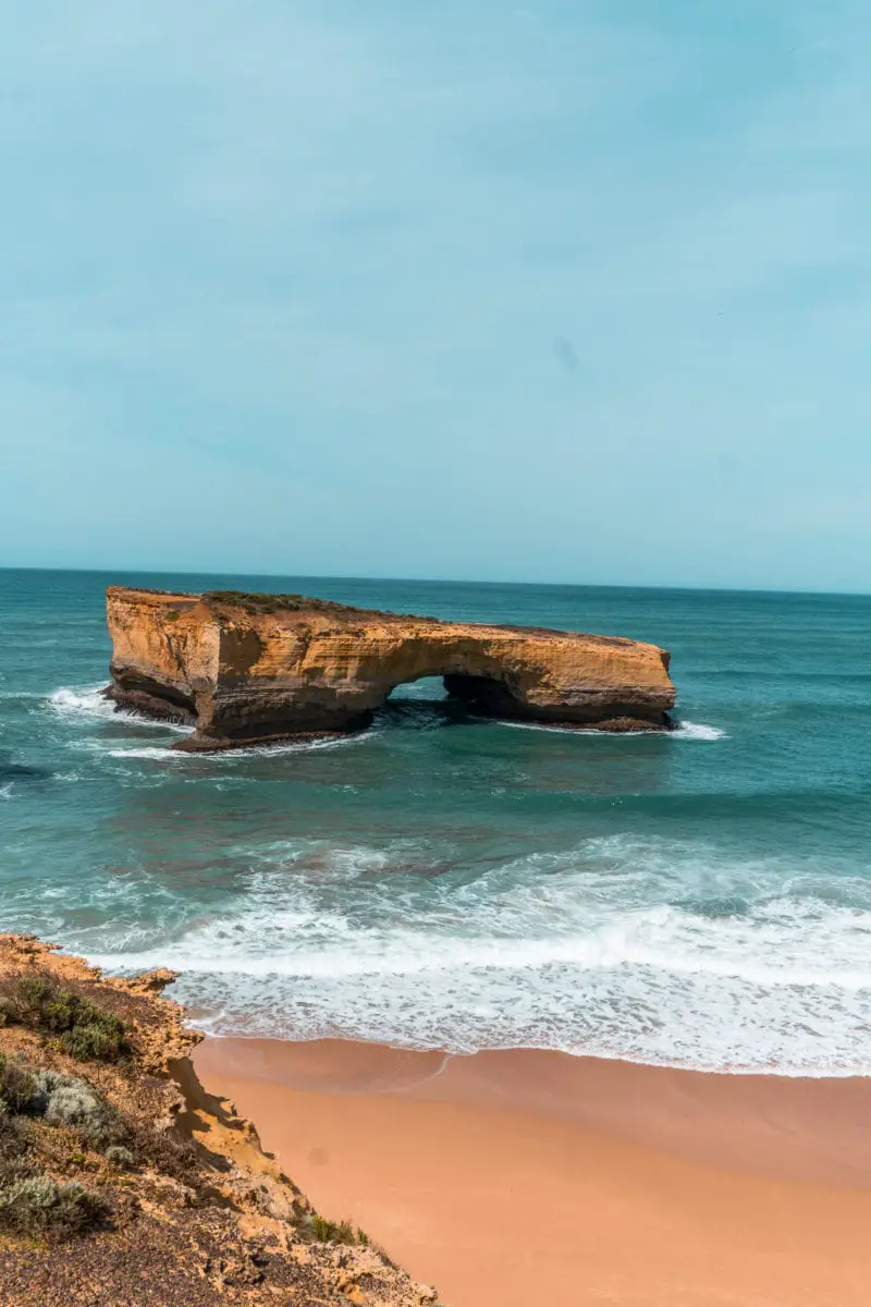 Natural rock formation resembling a bridge, known as London Bridge, standing in the sea near a sandy beach under a blue sky. London Bridge is located along the Great Ocean Road.