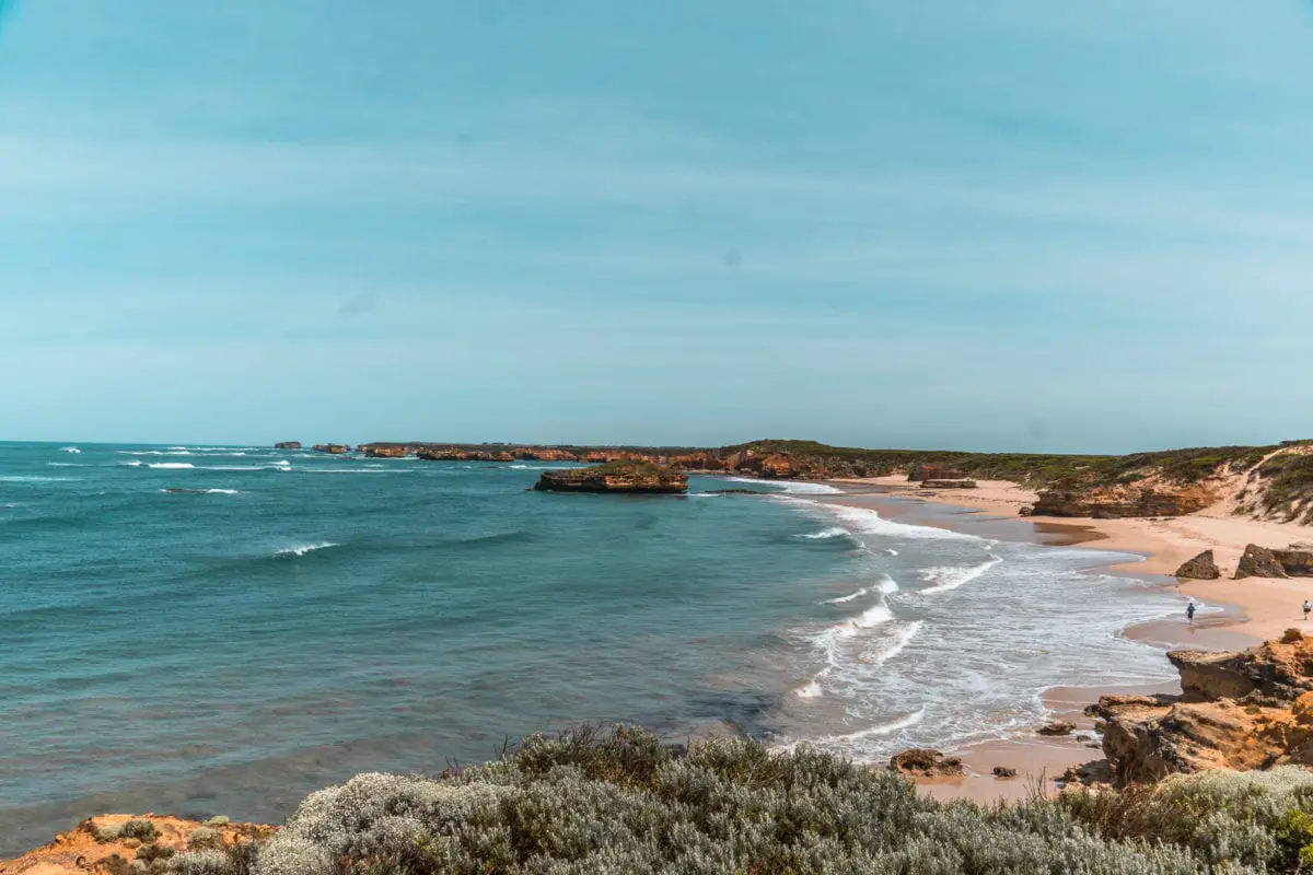 Panoramic view of a rugged coastline with waves washing onto the sandy shore, flanked by cliffs and vegetation under a clear sky. The Bay of Martyrs is an off the beaten path place to visit along the Great Ocean Road.