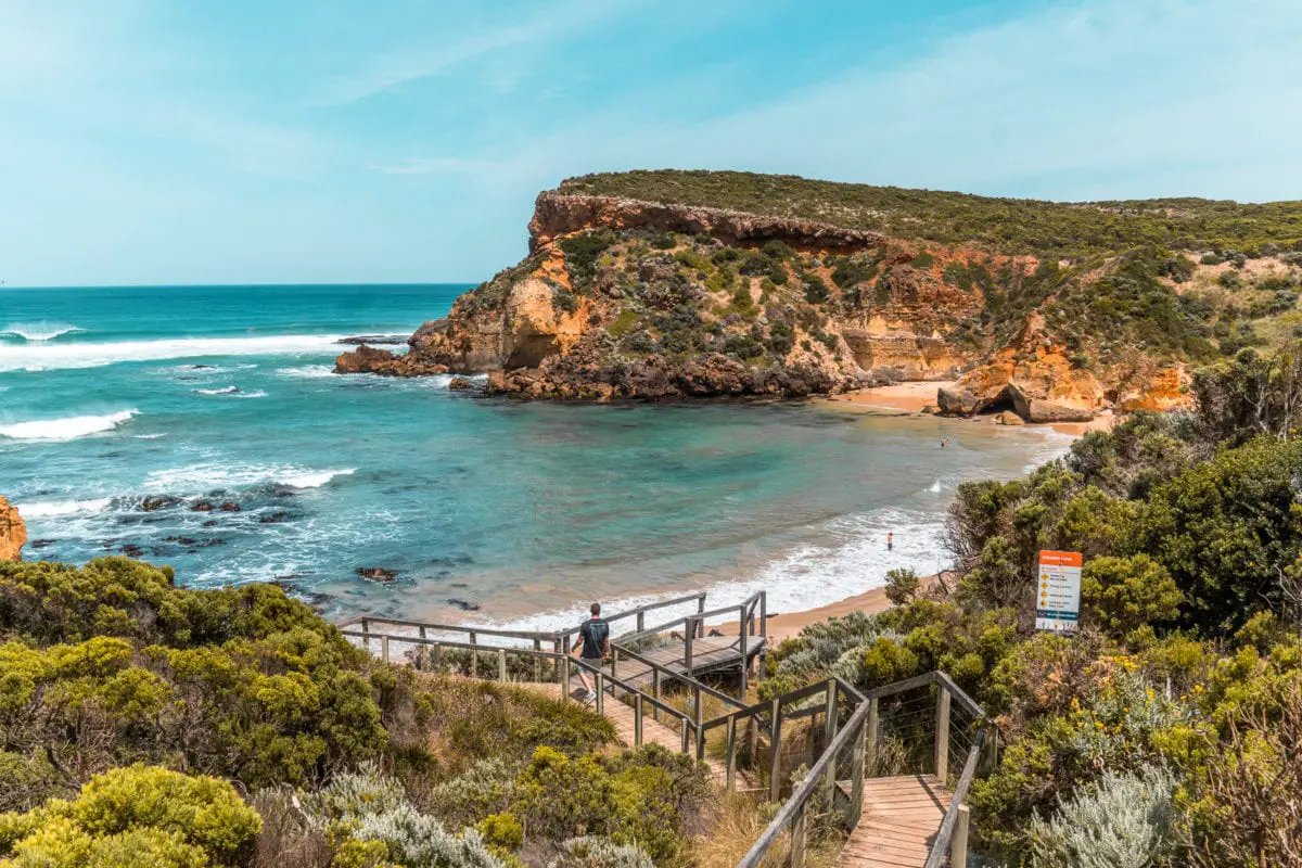 Wooden boardwalk leading to Childers Cove with waves crashing against the shore, surrounded by cliffs and greenery under a blue sky. Childers Cove is one of the not so secret beaches in Victoria - a must for your 3-day Great Ocean Road itinerary.