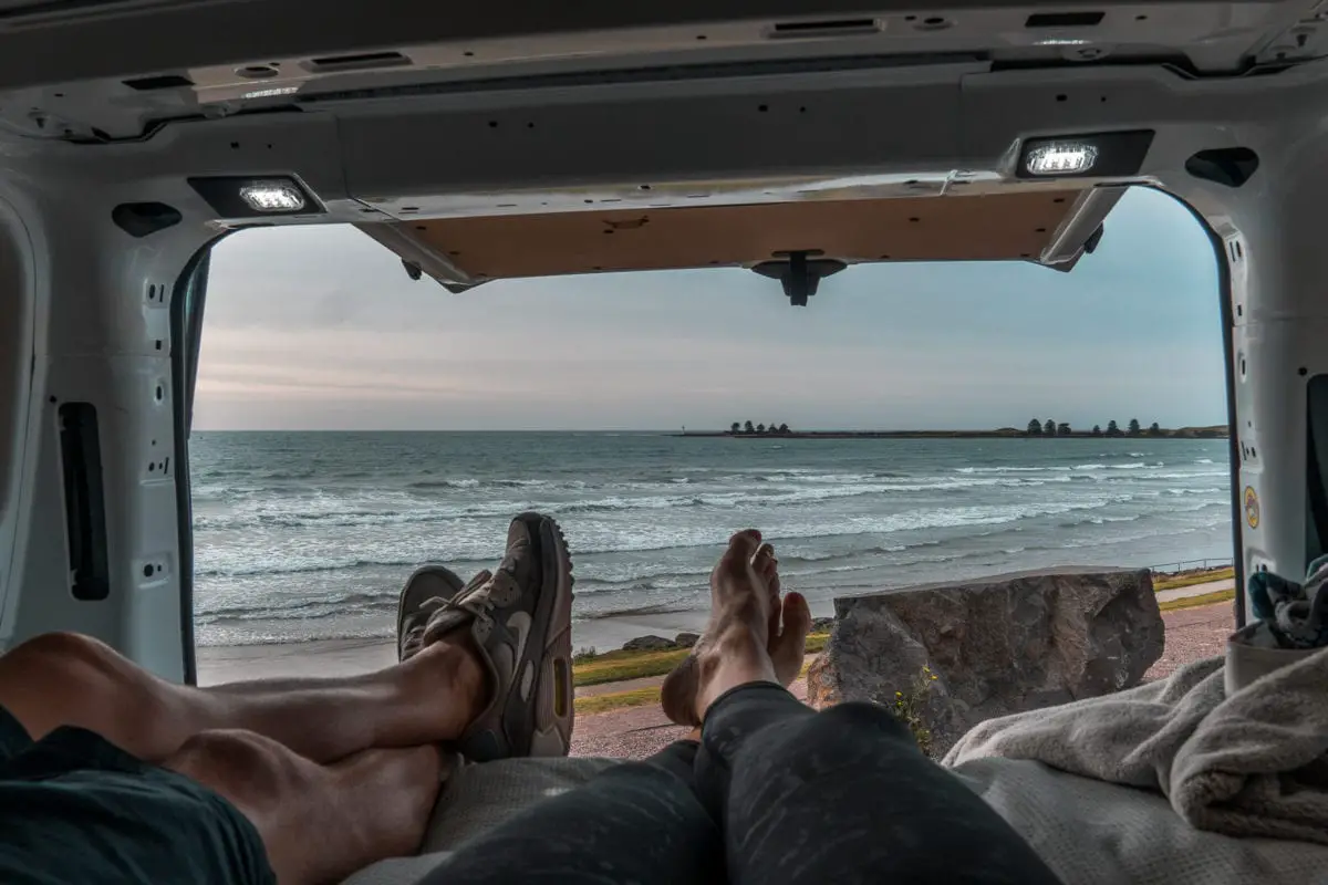 View from inside a vehicle with the rear door open, showing two legs stretched out towards the edge, with a beach and ocean in the background under a cloudy sky. The photo is taken in Port Fairy, Victoria, Australia.