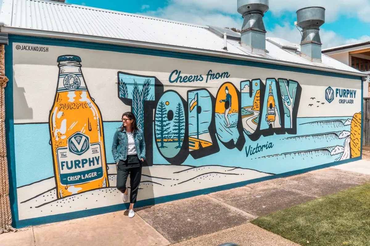 A person posing in front of a colorful mural with the text "Cheers from Torquay, Victoria" and an image of a beer bottle. Stop for a while in Torquay on your Great Ocean Road itinerary - this beachside town is where the road officially begins.