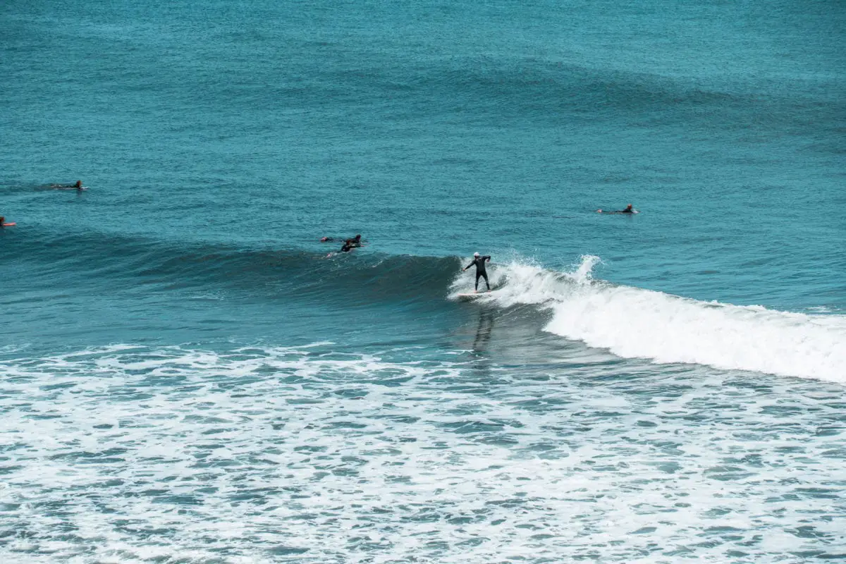 Surfers riding waves in the ocean, with one standing on a surfboard on a wave, and others paddling nearby. Bells Beach is one of the best beaches in Australia - and a must-visit on your Great Ocean Road trip.