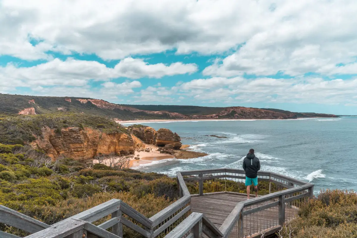 A person stands at a wooden lookout deck overlooking a sweeping coastal landscape with rugged cliffs and a sandy beach below a cloudy sky. Take a detour off the Great Ocean Road to visit Point Addis for the amazing views.