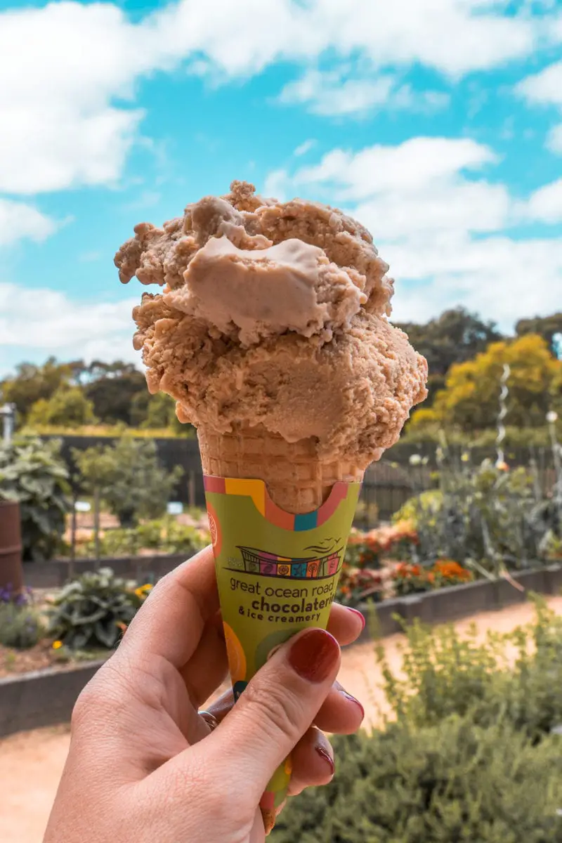 A hand holding up a large ice cream cone with a scoop of chocolate ice cream against a blurred background of a garden. Grab a snack from the Great Ocean Road Chocolaterie and Ice Creamery!