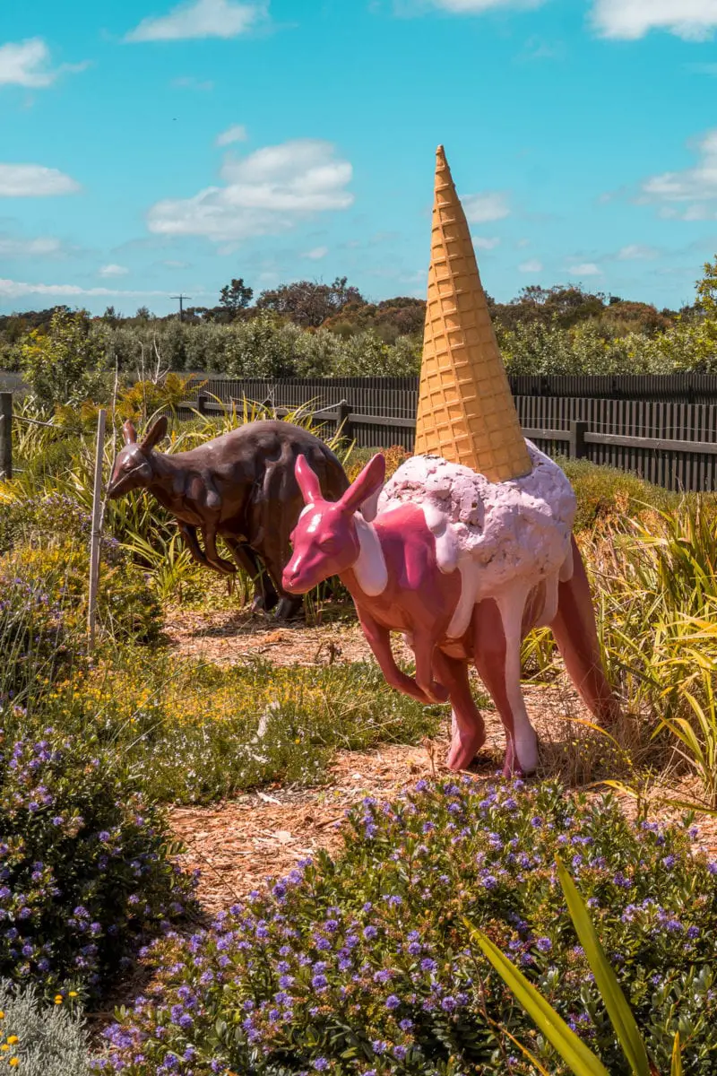 A whimsical sculpture of a kangaroo with a large ice cream cone on its back, placed among flowering plants and grasses. Grab a snack from the Great Ocean Road Chocolaterie and Ice Creamery!