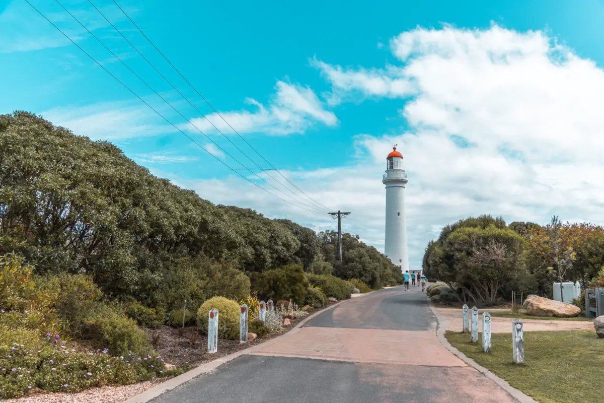 A road leading to a tall white lighthouse with a red cap, surrounded by greenery under a blue sky with clouds. Split Point Lighthouse in Aireys Inlet is one of the most iconic things to see on the Great Ocean Road.