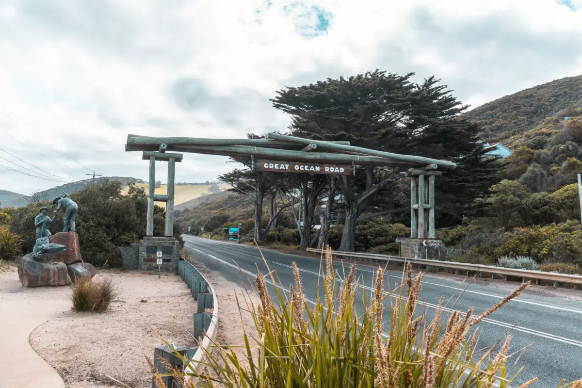 The entrance sign of the Great Ocean Road with a sculpture of two figures beside it, with the road stretching out ahead and a lush hillside in the background. One of the best photos along the Great Ocean Road is one of the Memorial Arch.