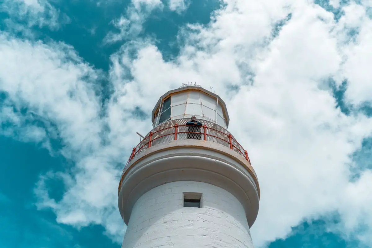 Looking up at a white lighthouse with a red railing around the lantern room, set against a backdrop of blue sky with scattered clouds. A man is standing on the platform looking into the distance. Climb the historic Cape Otway Lightstation for amazing views over the coastline.