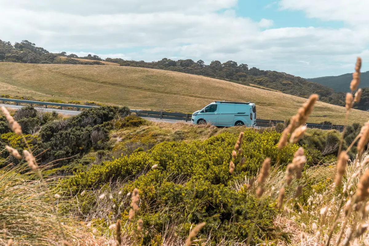 A white van driving on a coastal road with rolling hills in the background and vegetation in the foreground. Driving the Great Ocean Road is a trip of a lifetime - it's one of the best road trips in Australia.