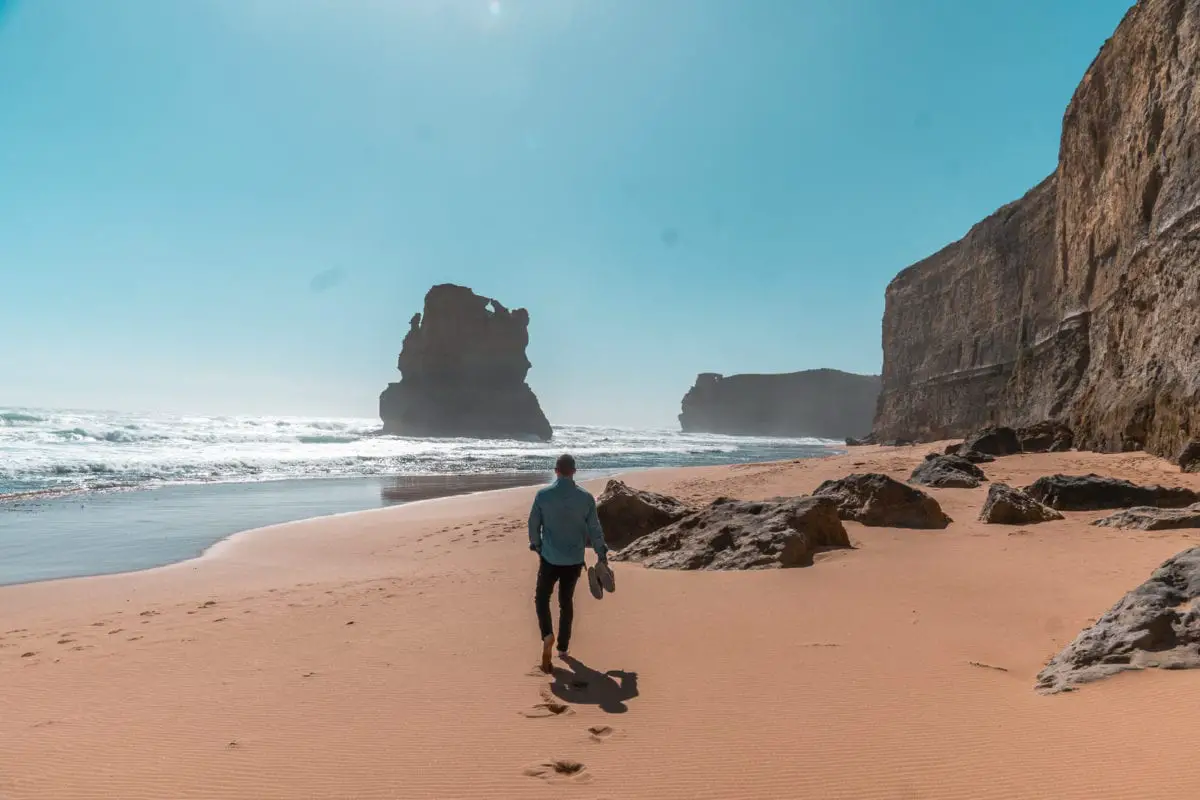 A man (the author's husband) walking towards a towering rock formation on a sandy beach, with waves crashing in the background and steep cliffs on either side under a blue sky. Make sure you stop by the Gibson Steps on the Great Ocean Road before visiting the 12 Apostles.