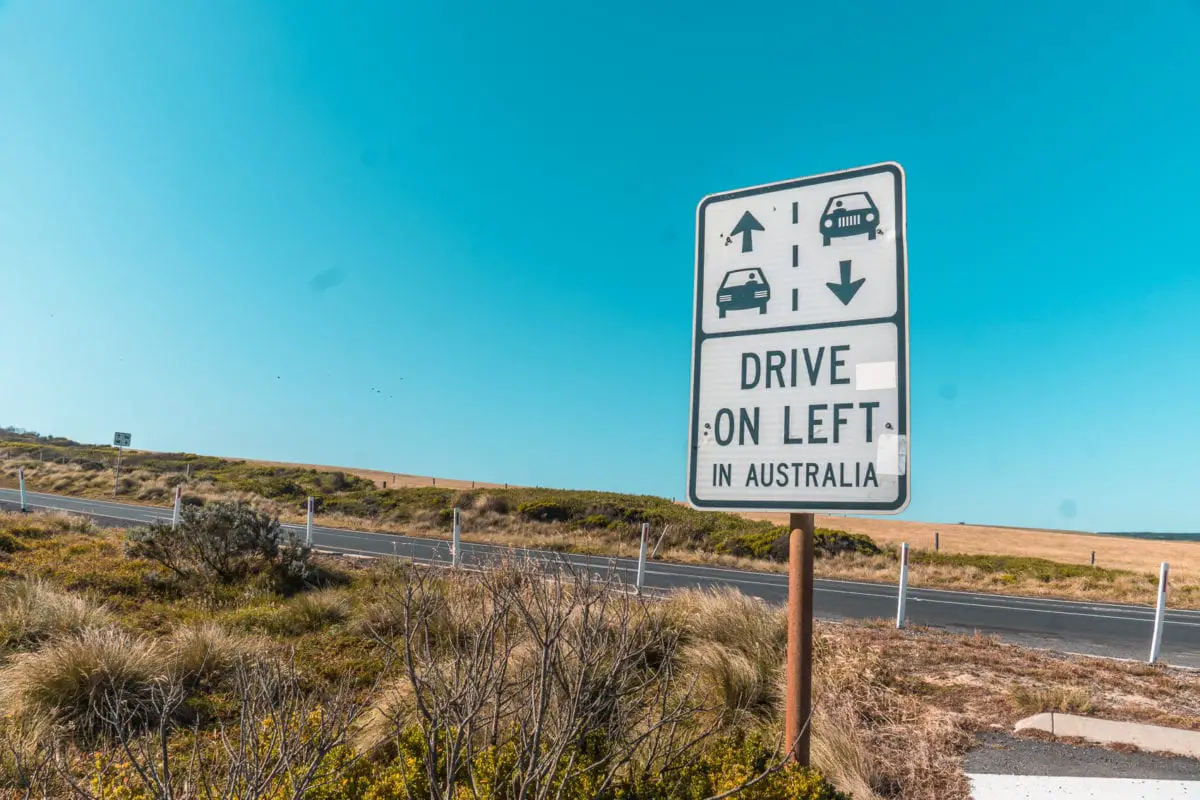 A road sign indicating "Drive on Left in Australia" with a blue sky in the background and a hint of the ocean in the distance. Make sure you drive on the left-hand side when you drive the Great Ocean Road in Australia!