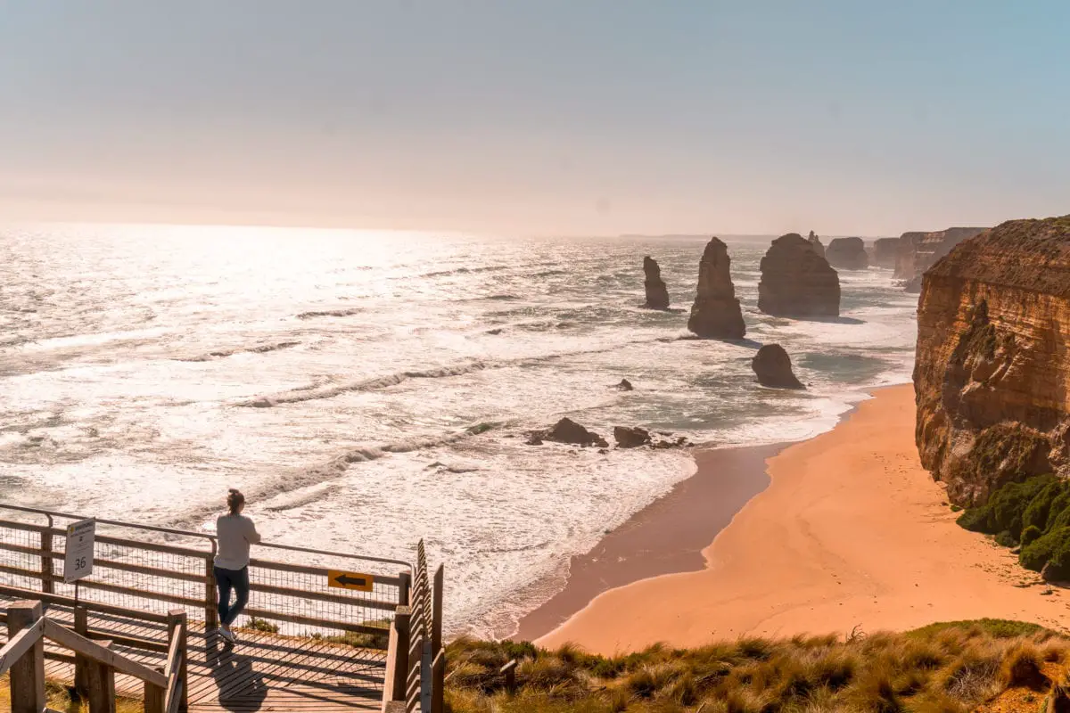 A woman (the author of the article) standing on a viewing platform overlooking a series of large rock stacks in the ocean known as the Twelve Apostles, with foamy waves and a sandy beach visible. The 12 Apostles are the most popular things to do on the Great Ocean Road - this will be the highlight of your trip!