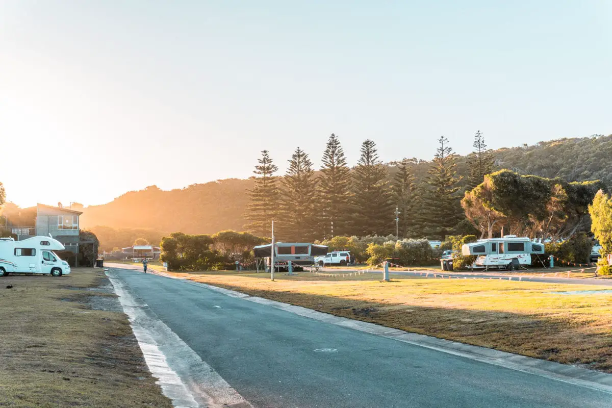 Caravans parked beside a coastal road at dusk, with trees and a soft glow from the setting sun in the background. Some of the best Great Ocean Road accommodation is at caravan parks and campground.