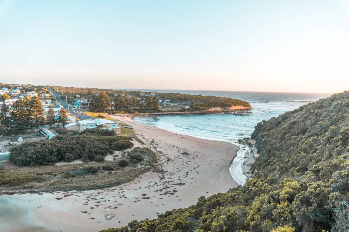 Aerial view of Port Campbell with buildings along the shoreline, a sandy beach, and a winding river entering the sea. Port Campbell is a great place to stay along the Great Ocean Road.