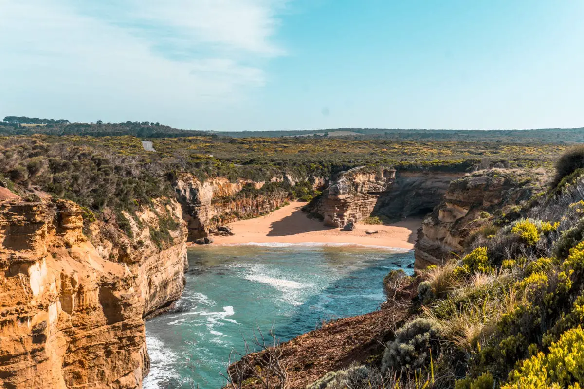 View overlooking a secluded beach cove with cliffs on either side and waves gently reaching the sandy shore. You must visit the Loch Ard Gorge, one of the best things to see on the Great Ocean Road.