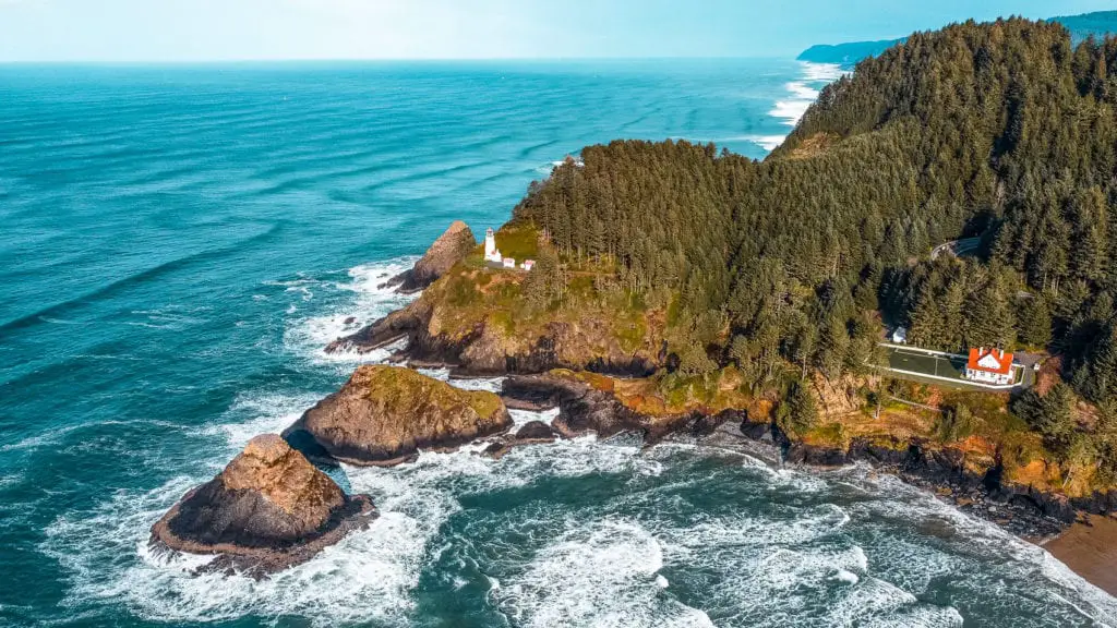 An aerial view of a rugged coastline with a historic lighthouse perched on a cliff, surrounded by dense forests and crashing waves below. The Oregon Coast is a stunning place to visit in the USA, offering wild coastlines and plenty of escapes to nature.
