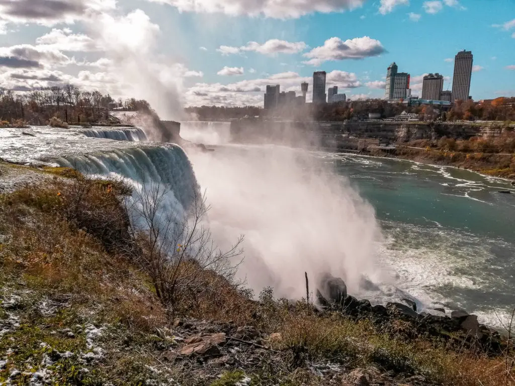 The misty spray of Niagara Falls with a backdrop of colorful autumn trees and distant city skyline under a cloudy sky. Niagara Falls will astound you with its beauty and power.