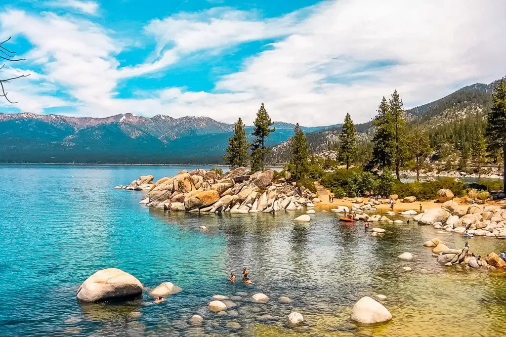 Tranquil waters of Lake Tahoe with smooth boulders along the shore, forested mountains in the distance, and a clear sky overhead. Lake Tahoe in California is a great destination to visit in the US year-round.