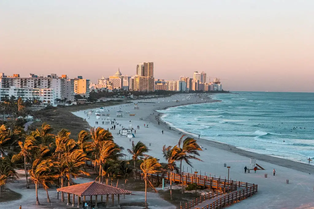 Miami Beach at sunset with soft pastel skies, the bustling beachfront filled with people, palm trees swaying in the breeze, and a view of the city's skyline in the distance. Miami is a great city to visit - there's beaches, city life and a lot of fun to be had.