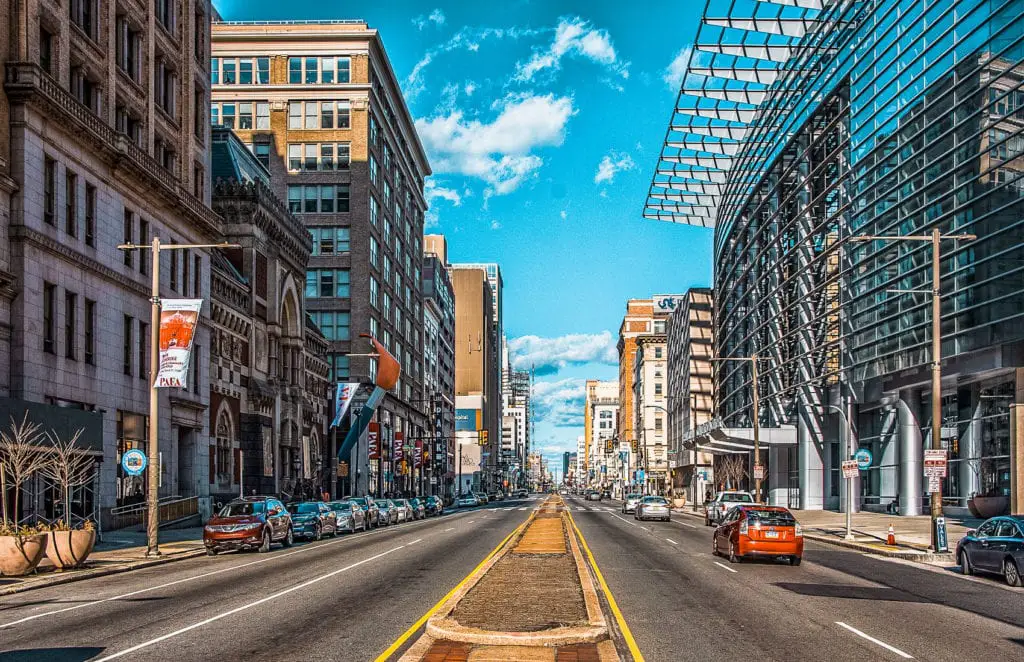Vibrant city street in the USA with a mix of traditional and modern architecture. Historic buildings with intricate facades stand beside a sleek glass structure under a clear blue sky with scattered clouds. The road is marked with a yellow double line and flanked by parked cars on either side, capturing a typical urban scene. There are so many things to do in Philadelphia, a very cool city.