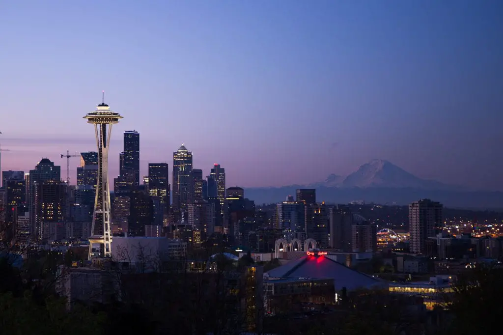 Dusk view of the Seattle skyline featuring the iconic Space Needle, city buildings illuminated against the twilight sky, and the silhouette of Mount Rainier in the distance. Seattle is one of the coolest cities to visit in the USA.