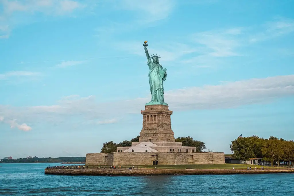 The iconic Statue of Liberty stands tall with a clear blue sky in the background, seen from the water's edge with visitors nearby. New York City is one of the most popular cities to visit in the USA.