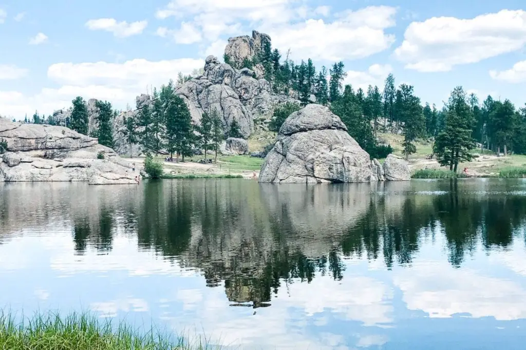 Sylvan Lake in Custer State Park, South Dakota, with impressive granite formations reflected in calm waters surrounded by green forest under a clear sky. There are so many things to do in South Dakota - it's one of the more underrated states to visit in the USA.