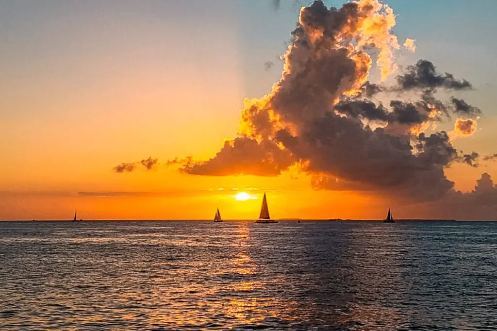 A breathtaking sunset at sea with vibrant orange hues in the sky, cloud formations, and silhouetted sailboats on the horizon. Watching the sunset is one of the best things to do in Key West, Florida.