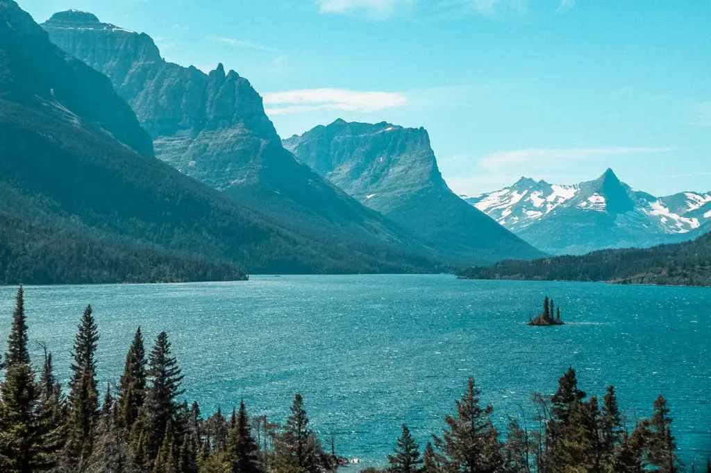 Scenic view of Saint Mary Lake in Glacier National Park with rugged mountain peaks, crystal-clear blue water, and a small island with tall trees. Stop by to explore one of the most stunning places in the USA - Glacier National Park.