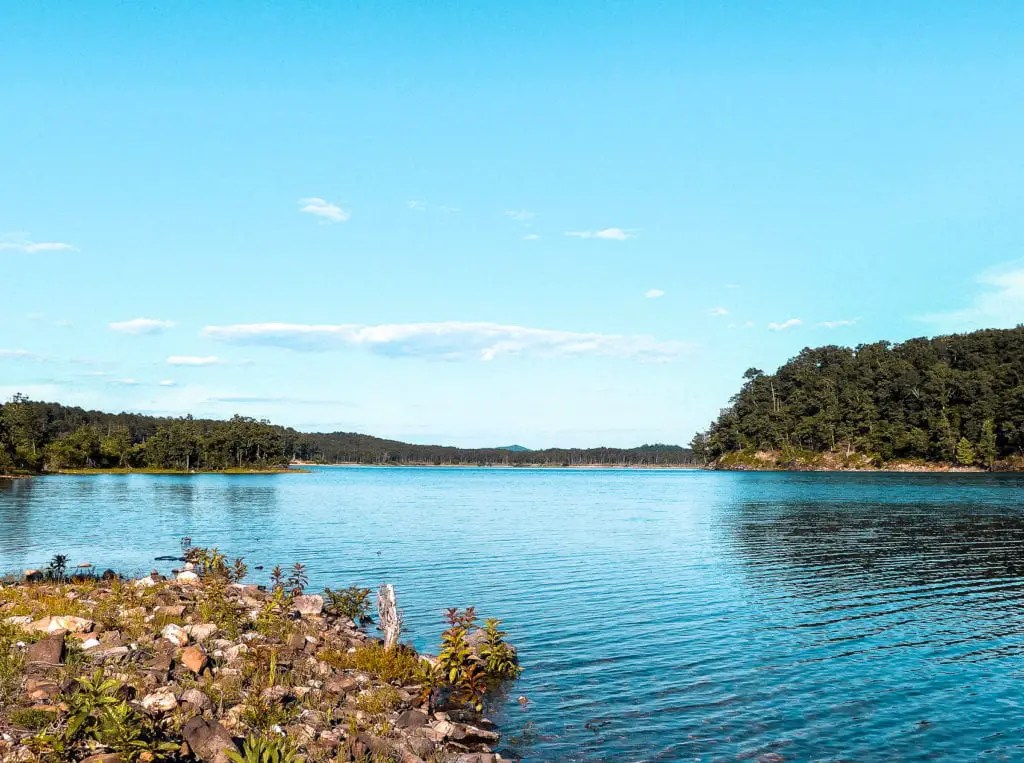 A serene lake with clear blue waters, bordered by lush greenery and distant rolling hills under a bright sky dotted with fluffy clouds.
