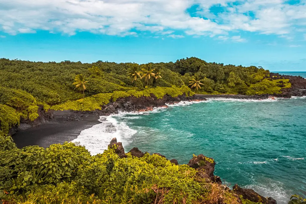 A serene black sand beach with lush tropical foliage on the shoreline, clear blue waters, and gentle waves in Hawaii. Waianapanapa Black Sand Beach on the Hawaiian island of Maui along Road to Hana, USA.