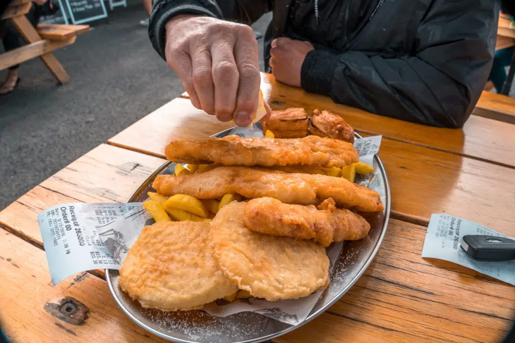 A person's hand squeezing a lemon over a large plate of fish and chips on a wooden table, outdoors. A must-do on any coastal trip in Australia is try the local fish and chips.