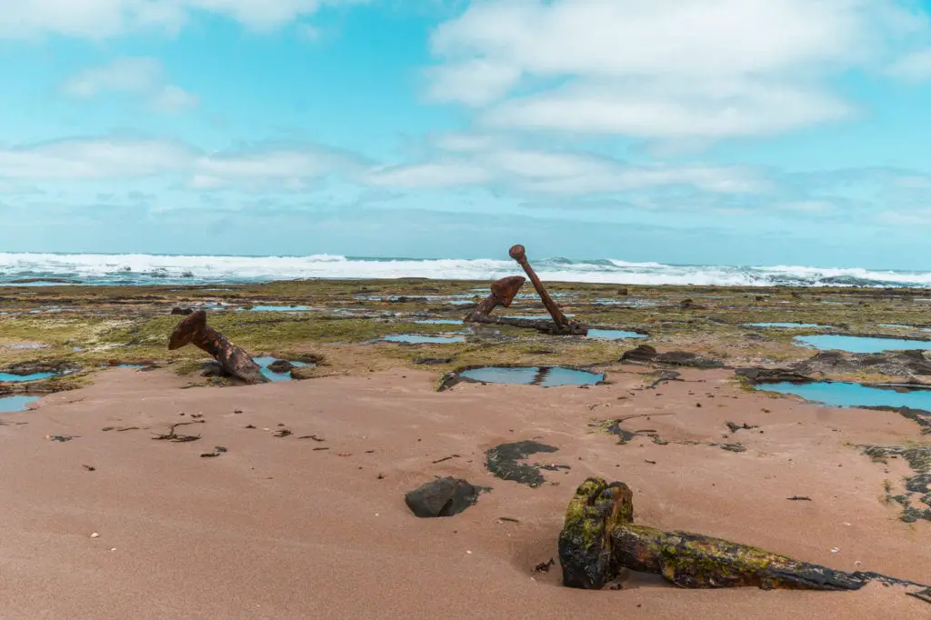 Rusty old anchors lying on a sandy beach with rock pools, ocean waves in the background under a cloudy sky. The coastline alongside the Great Ocean Road is known as the Shipwreck Coast, and it's easy to spot some of the wreckage from shipwrecks at Wreck Beach.