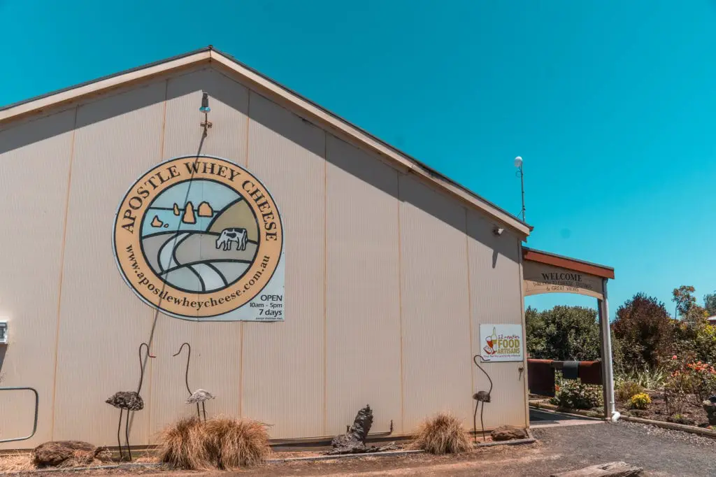 Exterior view of Apostle Whey Cheese building with a company logo, an open sign, and a "Welcome" archway, under a clear blue sky. Lush farmland sits alongside the Great Ocean Road, affording some great locally made produce. You can stop into farmhouses and producers along the 12 Apostles Gourmet Trail.