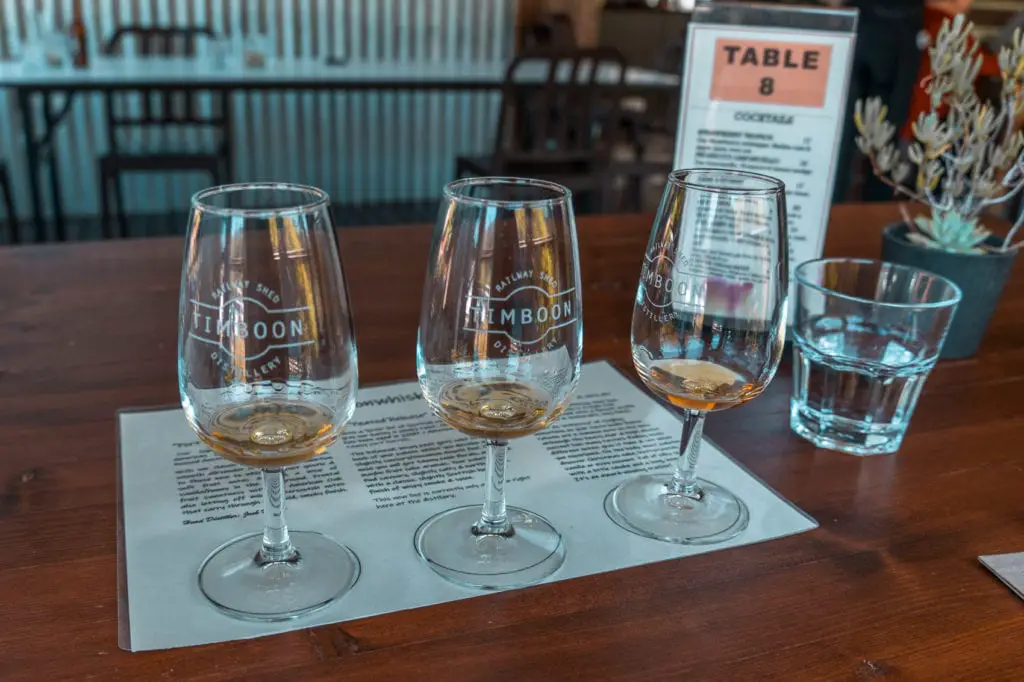 Three glasses of whiskey on a tasting mat with descriptions, on a wooden table inside a bar. Check out locally produced whisky and gin on your Great Ocean Road road trip.