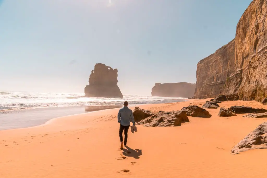 A person - the author's husband - walking on a sandy beach towards a large rock formation in the sea, with towering cliffs in the background. This is the Gibson Steps, one of the stops on the famous Great Ocean Road in Victoria, Australia.