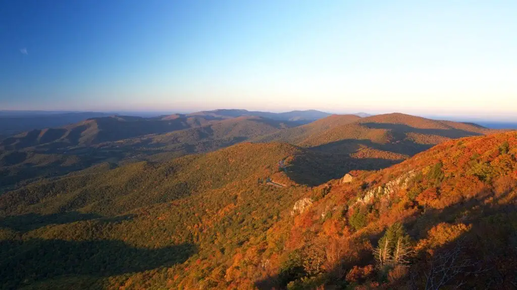 Expansive view from a mountain overlook at sunset, with shades of orange and red highlighting the fall foliage across rolling hills. This is the beautiful Shenandoah National Park.