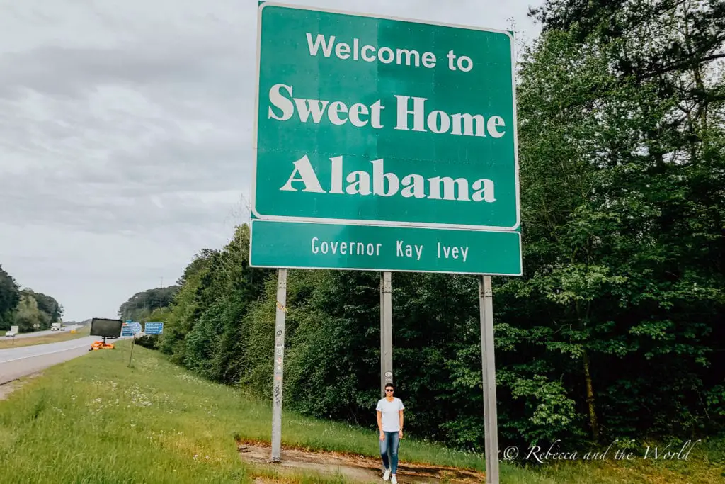 A person - the author of this article - standing beside a green highway sign reading "Welcome to Sweet Home Alabama," indicating the state border and offering a greeting to travellers. Stop in Alabama on your Deep South road trip - make sure to get a photo at the state border.