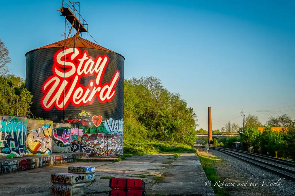 A large water tower with graffiti art that reads "Stay Weird" in bold red letters, accompanied by various other colorful tags. In the background, train tracks and industrial structures are visible.